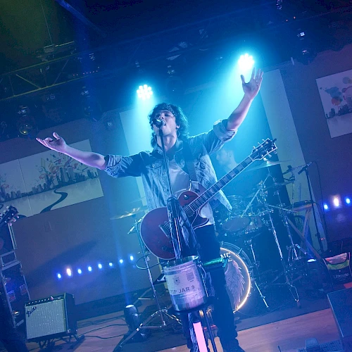 A band is performing on stage with blue lighting; one member has their arms raised, playing guitar, while others play bass and drums.
