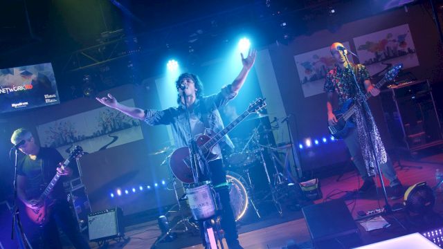 A band is performing on stage with blue lighting; one member has their arms raised, playing guitar, while others play bass and drums.