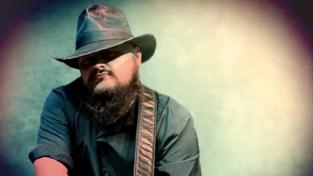 A person wearing a hat and holding a guitar, with a beard and dark clothing, set against a blurred, softly lit background.