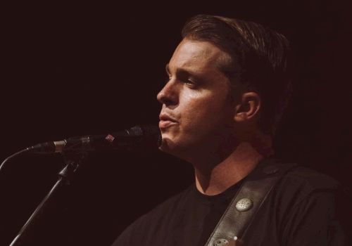 A person is playing an acoustic guitar and singing into a microphone on stage, wearing a black shirt against a dark background.