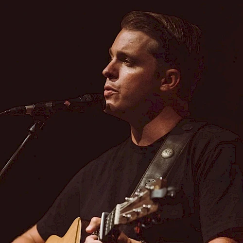 A person is playing an acoustic guitar and singing into a microphone on stage, wearing a black shirt against a dark background.