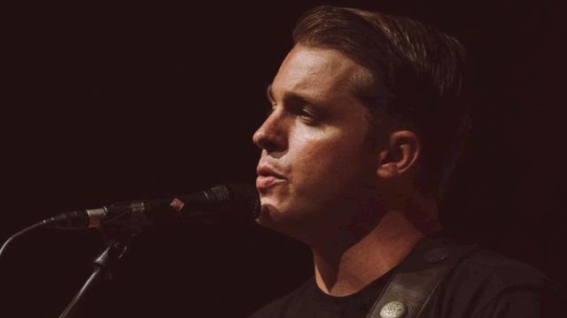 A person is playing an acoustic guitar and singing into a microphone on stage, wearing a black shirt against a dark background.