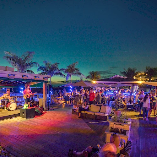 A lively outdoor beach bar scene with a band playing on stage, people enjoying the atmosphere, palm trees, and colorful lights at dusk.
