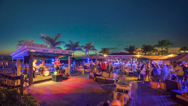 A lively outdoor beach bar scene with a band playing on stage, people enjoying the atmosphere, palm trees, and colorful lights at dusk.