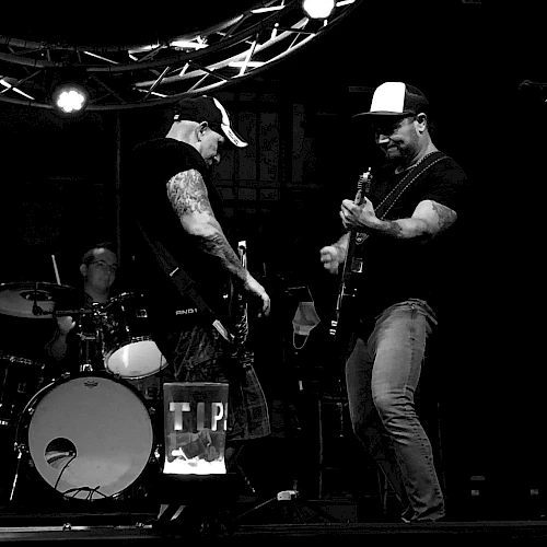 A black and white image of a band performing on stage, featuring two guitarists and a drummer under stage lights.
