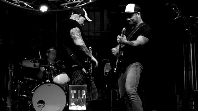 A black and white image of a band performing on stage, featuring two guitarists and a drummer under stage lights.