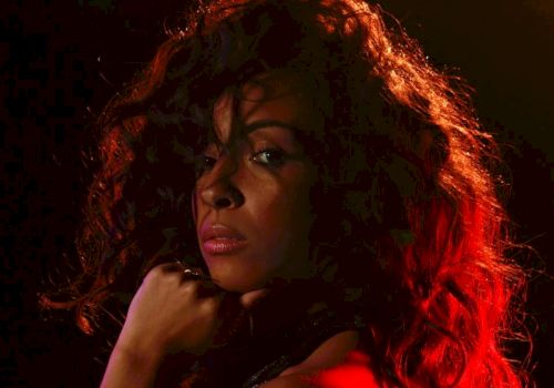The image shows a person with curly hair, illuminated with dramatic red and black lighting, looking towards the camera.