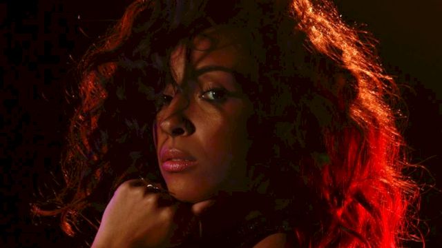 The image shows a person with curly hair, illuminated with dramatic red and black lighting, looking towards the camera.