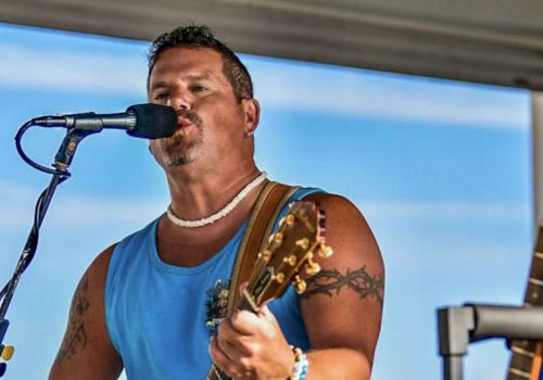A person is singing and playing guitar on stage, wearing a blue sleeveless shirt, with another guitar visible nearby.