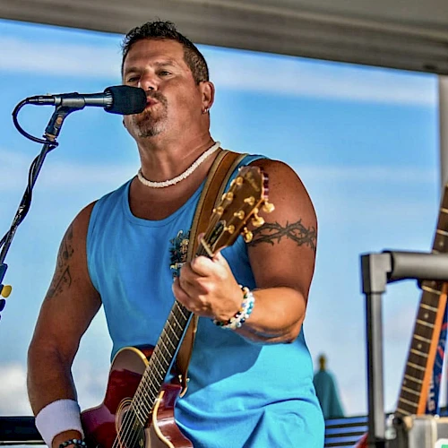A person is singing and playing guitar on stage, wearing a blue sleeveless shirt, with another guitar visible nearby.