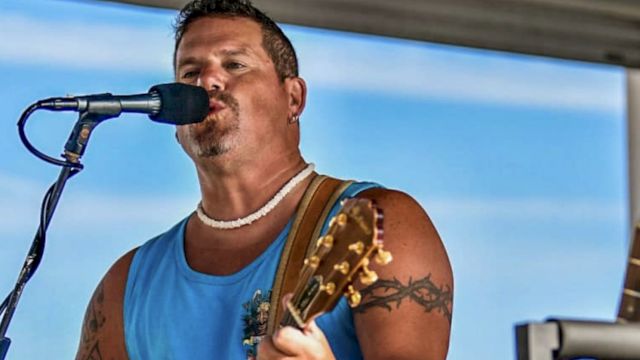 A person is singing and playing guitar on stage, wearing a blue sleeveless shirt, with another guitar visible nearby.