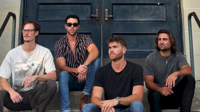 Four men sit on steps in front of a double door, wearing casual attire, with metal railings on either side.