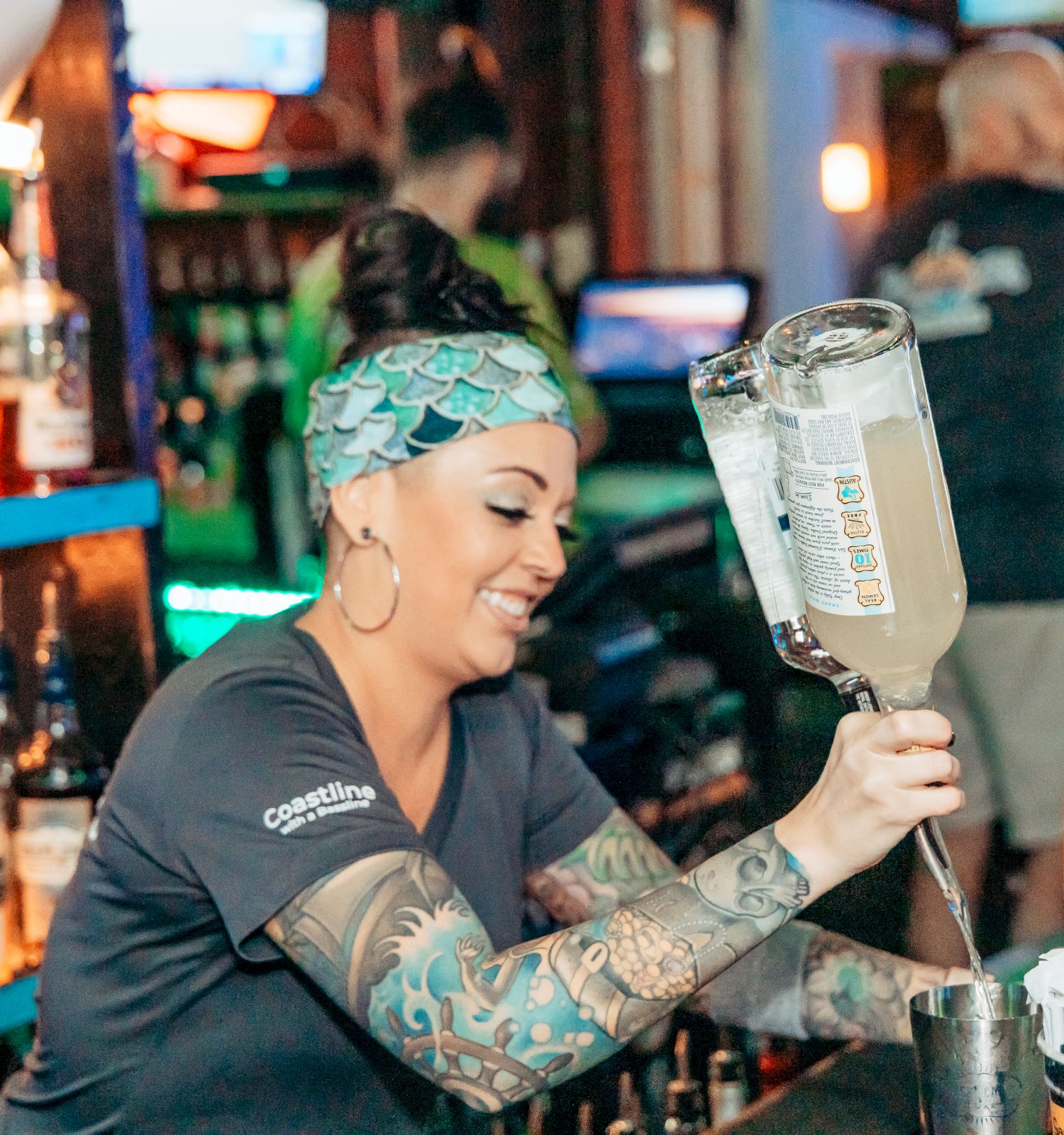 A bartender with tattoos and a headband pours a drink at a bar. Bottles and bar supplies are visible in the background.