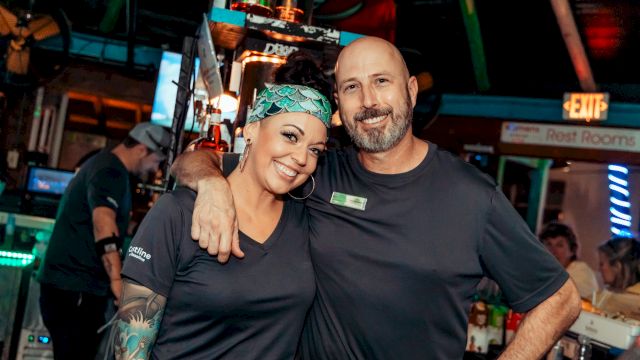 Two smiling coworkers pose arm-in-arm behind a bar, wearing black shirts; a tattooed woman with a bandana and a man with a beard.