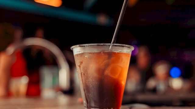A plastic cup with a dark beverage and ice, topped with a black straw, sits on a coaster in a dimly lit setting.