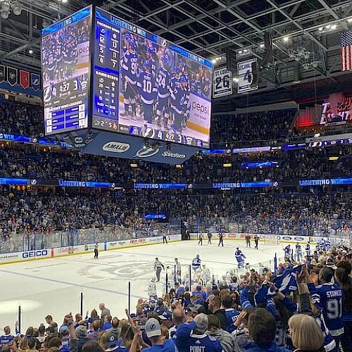 A large crowd watches a hockey game in an indoor arena with a big screen displaying the match and banners hanging above.