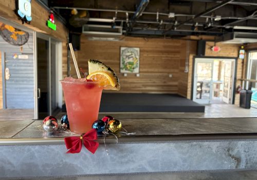 A tropical pink cocktail with a pineapple wedge and straw sits on a weathered bar counter, a red bow garland below, inside a rustic caf&eacute;.