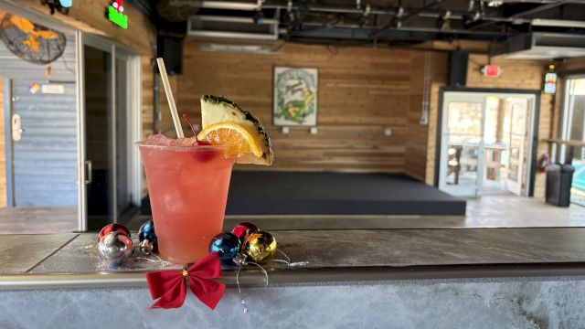A tropical pink cocktail with a pineapple wedge and straw sits on a weathered bar counter, a red bow garland below, inside a rustic café.