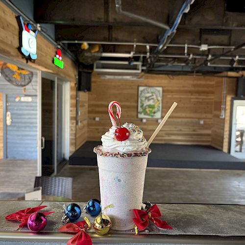 A creamy milkshake topped with cherries and sprinkles sits on a wooden bar, wrapped candies scattered around; cozy rustic interior behind it.
