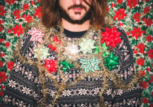 A bearded person wearing a patterned sweater wrapped in shiny garlands and oversized bows against a red-green floral backdrop.