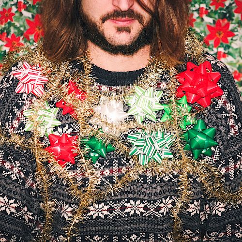 A bearded person wearing a patterned sweater wrapped in shiny garlands and oversized bows against a red-green floral backdrop.