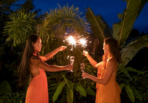 Two women in peach dresses toast sparklers outdoors at night, flames bright, tropical plants backdrop, festive and joyful moment.