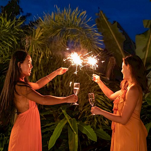 Two women in peach dresses toast sparklers outdoors at night, flames bright, tropical plants backdrop, festive and joyful moment.