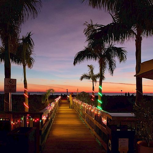 Sunset at a boardwalk leading to the sea, palm trees lit with colorful lights, warm evening glow on the horizon.