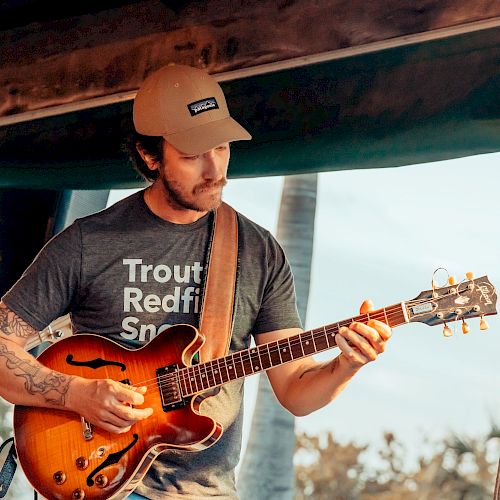 A guitarist wearing a tan cap and grey T-shirt plays a sunburst hollow-body guitar on a wooden stage outdoors, focused on the fingers.