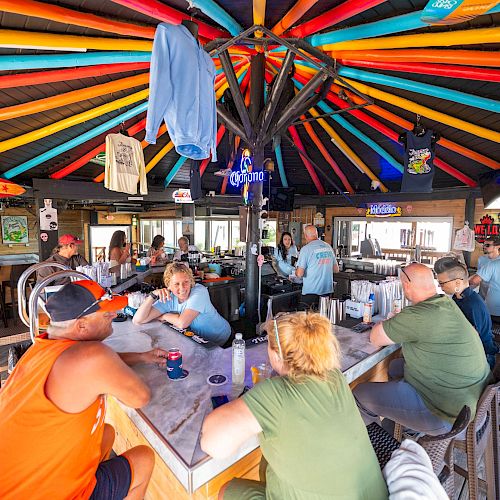 Group of people dining inside a colorful, sunburst-roofed shack with long tables, drinks, and casual conversation. A laid-back, beachy bar vibe.