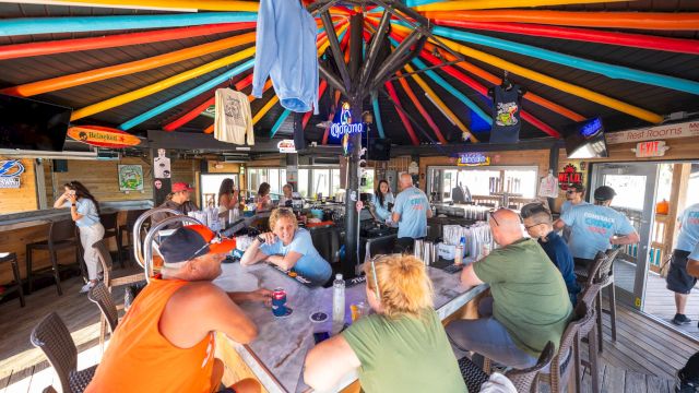Group of people dining inside a colorful, sunburst-roofed shack with long tables, drinks, and casual conversation. A laid-back, beachy bar vibe.