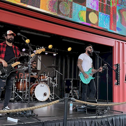 A four-member rock band performs on an outdoor stage, with guitars, drums, a bassist, and a female singer under a colorful backdrop.
