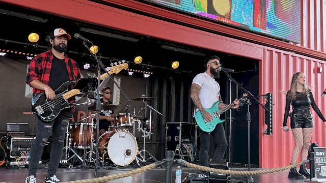 A four-member rock band performs on an outdoor stage, with guitars, drums, a bassist, and a female singer under a colorful backdrop.