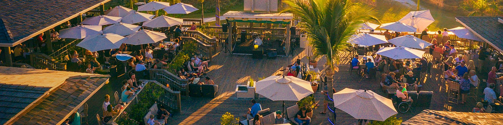 Beachfront party scene at sunset — palm trees, umbrellas, and a crowd mingling by a beachfront venue. Enjoying drinks, music, and ocean views.