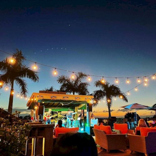 A beachside lounge at dusk with string lights, palm trees, and a small stage or bar area; colorful couches, people socializing.