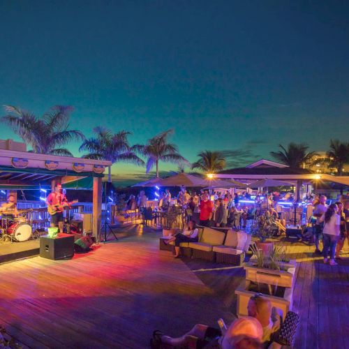 A lively outdoor beach party at dusk with a live band, lenient lighting, palm trees, sunbeds, and groups mingling on a wooden deck.