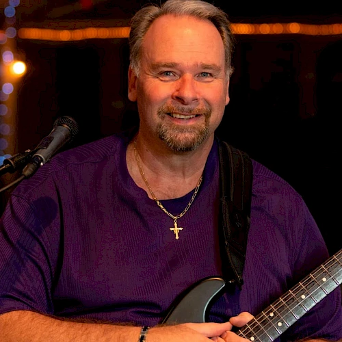 A smiling man with a purple shirt and guitar, wearing a necklace with a cross, on stage with warm lights.