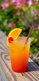 A colorful tropical drink in a tall cup with orange-to-red gradient, garnished with an orange slice and cherry, on a wooden railing.