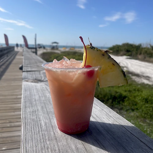 A tropical pink drink with ice on a weathered wooden railing, a pineapple wedge garnish, beachy sky and boardwalk in the background.