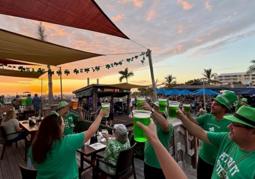 A group of people in green shirts and hats toast with bright green drinks on a beachside deck at sunset, festive celebration under canopies.