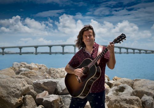 A man with a guitar stands on rocks by the water, a long bridge in the background under a blue sky with fluffy clouds.