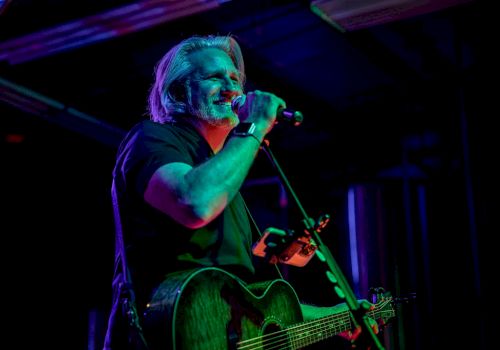 A male musician with a guitar sings into a microphone on a dark, colorful stage.