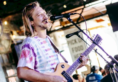 A man with long hair plays a light-colored electric guitar on stage, singing into a microphone during a live outdoor performance.