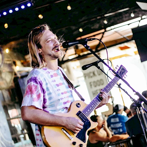 A man with long hair plays a light-colored electric guitar on stage, singing into a microphone during a live outdoor performance.