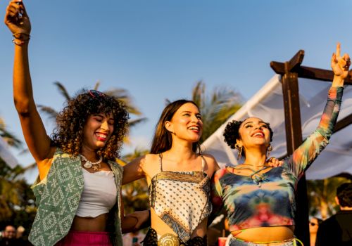 Three friends celebrate at an outdoor event, raising their hands with smiles as the sun shines, colorful outfits and a festive vibe.