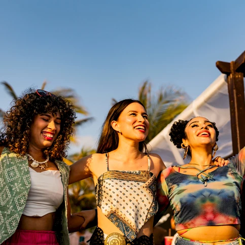 Three friends celebrate at an outdoor event, raising their hands with smiles as the sun shines, colorful outfits and a festive vibe.