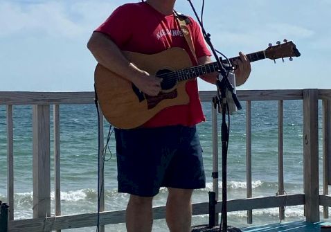 A man in a red shirt and cap plays an acoustic guitar on a seaside stage, singing into a mic with the ocean and railing in the background.