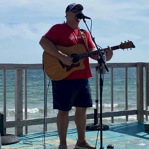 A man in a red shirt and cap plays an acoustic guitar on a seaside stage, singing into a mic with the ocean and railing in the background.