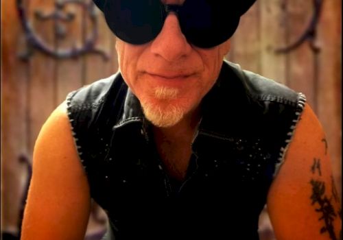 A man wearing a black hat, large sunglasses, and a sleeveless vest with tattoos on his arm, posing outdoors with a wrought-iron gate in the background.