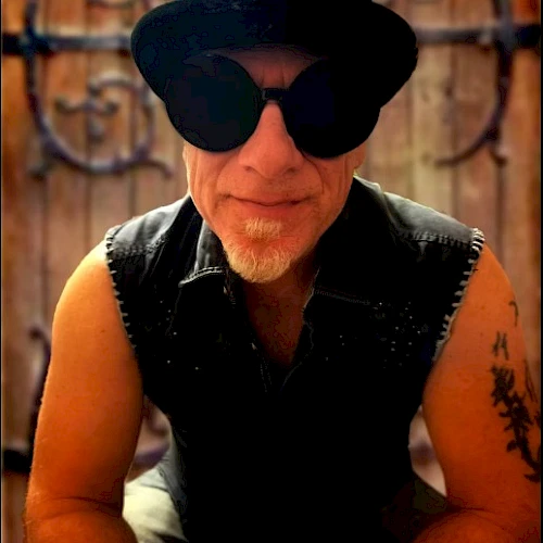 A man wearing a black hat, large sunglasses, and a sleeveless vest with tattoos on his arm, posing outdoors with a wrought-iron gate in the background.